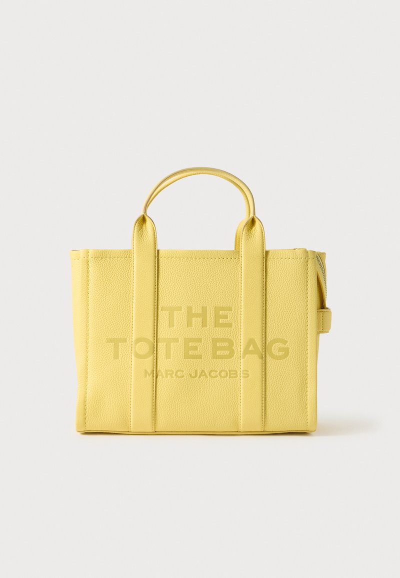 Žlutá kožená taška s dvojitými rukojeťmi, na které je embosovaný nápis "THE TOTE BAG" a "MARC JACOBS". Hladký povrch, strukturovaný tvar.