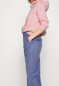 Sudadera rosa con bolsillo tipo canguro, que cuenta con capucha con cordón. Combinada con pantalones cargo azul claro con grandes bolsillos laterales y detalles de costura.