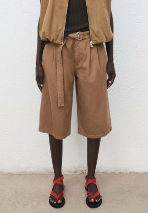 Person iført brune knælange shorts med bælte, en brun top og røde åbne sandaler, står op ad en hvid væg.