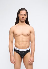 Άνδρας με μακριά πλεξούδα, φοράει μαύρα εσώρουχα Calvin Klein με λευκή μέση και λογότυπο, στέκεται μπροστά σε απλό φωτεινό φόντο.