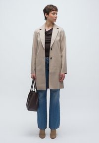 Μπεζ παλτό από μείγμα μαλλιού με γιακά με εγκοπές και δύο κουμπιά, σετ με μπλε τζιν με φαρδύ πόδι, καφέ τοπ και μποτάκια ταμπά.