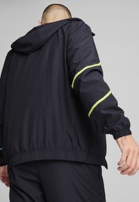 Giubbotto a vento blu navale con cappuccio e polsini elasticizzati, con accenti verde brillante lungo le maniche e orlo stringato.