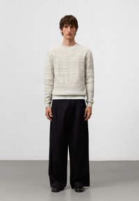 Jonge man die staat met een lichtgrijze trui, wit onderhemd, ruime zwarte broek en zwarte schoenen voor een effen witte achtergrond.