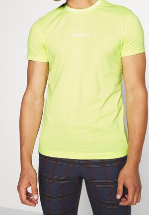 Homme portant un t-shirt jaune vif Calvin Klein Jeans et un pantalon à carreaux foncés, debout avec les bras détendus le long du corps.