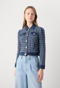 Veste cropped en tweed bleu avec des accents en denim, quatre poches frontales et des fermetures à boutons en argent. Manches longues avec poignets côtelés.
