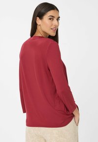 Mujer con cabello largo y oscuro, vistiendo una blusa roja de manga larga y pantalones beige con estampado, mirando por encima del hombro con las manos en los bolsillos.