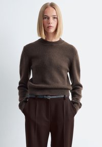 Frau mit blonden Bob-Haaren, die einen braunen Strickpullover und dunkle braune Hosen trägt und mit den Händen in den Taschen vor einem schlichten weißen Hintergrund steht.