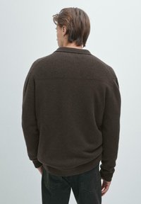 Muž čelem k odrazu, oblečený v tmavě hnědém žebrovaném svetru a černých džínách, na pozadí v jednobarevné světle šedé barvě.