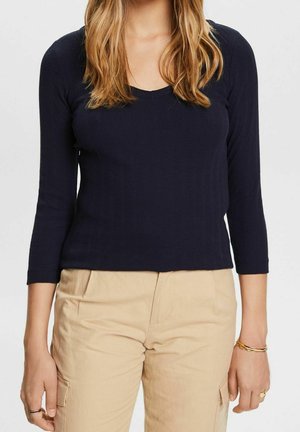 Woman wearing a navy blue long-sleeve top and beige cargo pants, with gold bracelets on right wrist and rings on left hand.