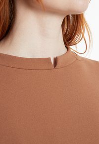 Braunes, strukturiertes Oberteil mit hohem Halsausschnitt und einem kleinen Schlüssellochdetail am Kragen. Glatte Stoffe mit dezentem Glanz.