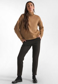 Brauner Rollkragenpullover aus weichem Strick, kombiniert mit dunkel gestreiften, taillierten Hosen. Das Model trägt schwarze, glänzende Loafer. Einfaches, modernes Outfit.