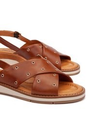 Sandalias de cuero marrón con tiras entrecruzadas que presentan ojetes de metal. Plantilla suave y acolchada, suela de cuña con un acento de raya blanca.