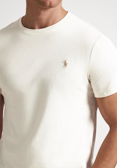 T-shirt en coton couleur crème avec col rond, manches courtes et petit logo brodé sur la poitrine. Texture douce, coupe classique.