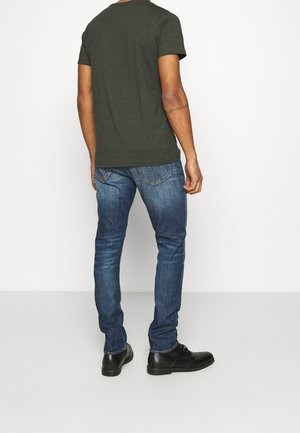 Mørkegrøn T-shirt med korte ærmer, blå slim-fit jeans og sorte sko. Synlige sømme og baglommer på jeans. Enkel, afslappet påklædning.
