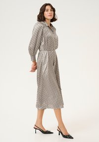 Grijze jurk met stippen, lange mouwen, knoopsluiting bij de kraag, elastische tailleband en halverwege de kuit; gecombineerd met zwarte slingback hakken.
