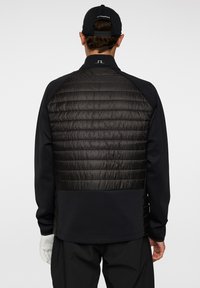 Person som bär en svart, vadderad, atletisk jacka med hög krage och handske, sedd bakifrån mot en enkel vit bakgrund.