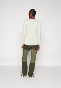 Svetlý dlhý rukáv top v off-white látke, v kombinácii so zelenými cargo nohavicami s vreckami a bočnými gombíkmi, viditeľné zozadu.