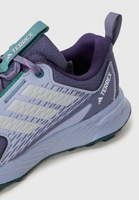 Baskets de sport violettes et bleu clair avec une tige en maille texturée, un bout renforcé et des accents verts, présentant trois bandes blanches sur le côté.