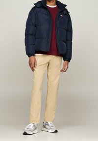 Navy pufferjacka med dragkedja, elastiska ärmar, röd och vit logotyp. Burgunder sweatshirt, beige byxor, grå och vita sneakers med nätdetaljer.