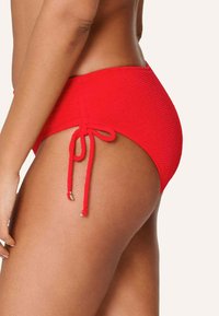 Červené plavky s vysokým pasem vyrobené z texturované, žebrované látky. Mají boční šňůrku se metalickými akcenty na koncích.