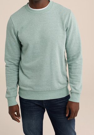 Mężczyzna w jasnozielonej bluzie z okrągłym dekoltem i ciemnoniebieskich dżinsach, stojący z ramionami luźno opuszczonymi na beżowym tle.