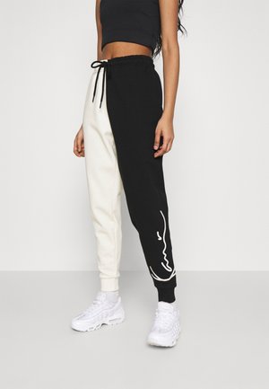 Kobieta w czarno-białych podzielonych spodniach dresowych ze sznurkiem w talii, czarnym krótkim topie i białych sneakersach na jednolitym tle.