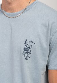 Camiseta de algodón color azul claro con un gráfico de una garza azul oscuro rodeada de hojas verdes en el lado izquierdo del pecho. Mangas cortas.