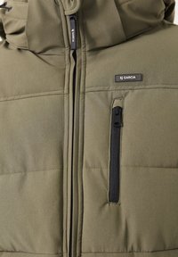 Chaqueta aislante verde oliva con textura acolchada, que presenta un cierre frontal con cremallera, bolsillos laterales y una etiqueta con el logo en el pecho.