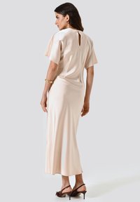 Abito in satin rosa pallido con schiena a fessura, maniche corte e una silhouette fluida; abbinato a sandali con tacco marroni.