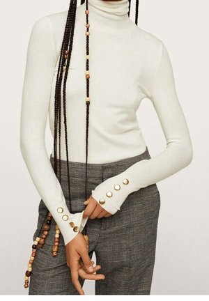 Biała, długi rękaw golf z złotymi guzikami na mankietach, zestawiony z szarymi, wysokim stanem spodniami w kratę oraz długimi zaplecionymi włosami z drewnianymi koralikami.