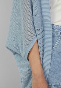 Lichtblauwe gebreide cardigan met open geweven design, geribbelde rand en korte mouwen. Draagt over een witte top, gecombineerd met denim.