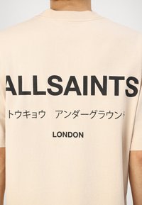 Beige cotton t-shirt featuring large black text that reads "ALLSAINTS" and "LONDON" with Japanese characters underneath. Plain design, short sleeves.
