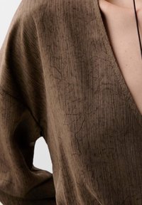 Top suelto de color marrón con textura, escote en V profundo y patrón de canalé. La tela suave se adapta suavemente, revelando parte del hombro.