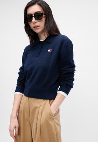 Námornícky modrý pletený polo sveter s rebrovaným lemom a manžetami, s malou logovou nášivkou. V kombinácii s béžovými nohavicami s vysokým pásom.