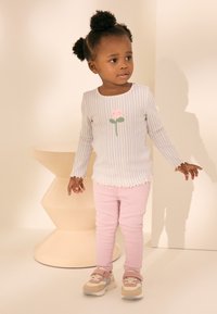 Lichtblauw-wit gestreept shirt met lange mouwen en een bloemmotief, gecombineerd met roze legging en multikleuren sneakers, staand bij een lichtgekleurde voetstuk.