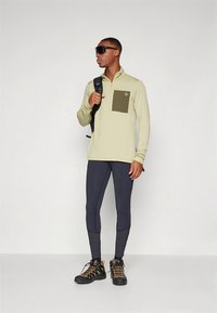 Lys beige langermet skjorte med olivengrønn brystlomme, kombinert med mørke leggings og svarte tursko. Ryggsekk og solbriller.