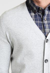 Světlá šedá cardigan s výstřihem do V, měkkou strukturou a tmavými knoflíky. Pod ním je modrá košile s kostkovaným vzorem a knoflíky.