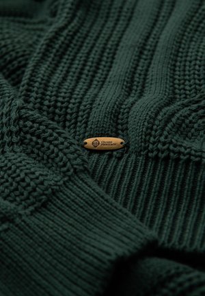Maglione lavorato a maglia verde scuro con texture a coste e motivi in contrasto. Presenta un piccolo tag in legno con la scritta "Ombre Heritage" incisa.