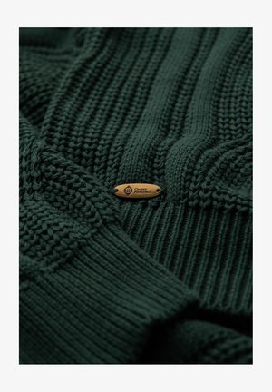 Maglione lavorato a maglia verde scuro con texture a coste e motivi in contrasto. Presenta un piccolo tag in legno con la scritta "Ombre Heritage" incisa.