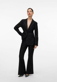 Blazer noir texturé avec un décolleté en V profond, associé à un pantalon évasé assorti. Les accessoires comprennent des talons aiguilles avec un motif contrastant.