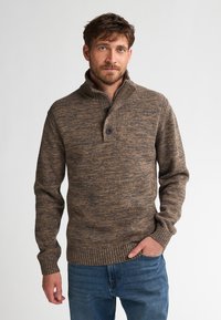 Mężczyzna w brązowym dżersejowym swetrze z zapięciem na guziki i niebieskich dżinsach, stojący na tle jasnego, jednolitego tła.