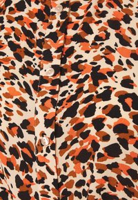 Tessuto con stampa leopardata in beige, caratterizzato da grandi macchie in nero, marrone e arancione. Quattro bottoni chiari decorano la parte anteriore.