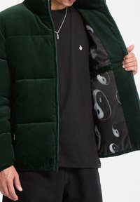 Green corduroy puffer jacket with quilted sections. Interior features black lining with a silver yin-yang pattern. Zipper and soft texture.