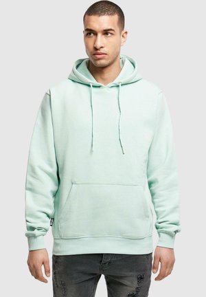Hoodie vert menthe en tissu doux, avec poche kangourou, capuche à cordon de serrage et poignets côtelés. Porté avec un jean gris usé.