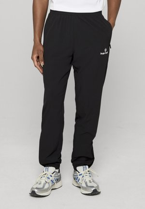 Mann iført svarte Sergio Tacchini treningsbukser og hvite og blå løpesko, stående med en hånd i lommen mot grå bakgrunn.