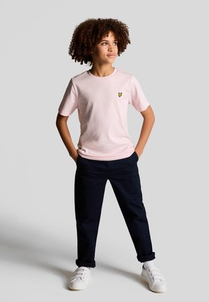 Dijete s kovrčavom kosom stoji s rukama u džepovima, nosi svijetloružičastu majicu, tamne hlače i bijele tenisice na jednobojnoj pozadini.