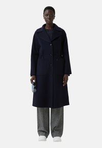 Marineblauwe wollen jas met een brede kraag, dubbele knopen aan de voorkant en twee voorzakken. Model houdt een zonnebril vast, draagt een broek met een patroon en witte schoenen.