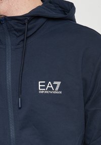 Felpa zip con cappuccio di colore blu navy realizzata in un tessuto morbido, con un logo bianco sul davanti. Include un cappuccio regolabile e una zip in contrasto.