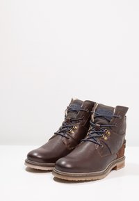 Braune Leder Stiefel in Knöchelhöhe mit einer glatten Textur, blauen Schnürsenkeln, Metallösen und einem kontrastierenden braunen Akzent an der Ferse.