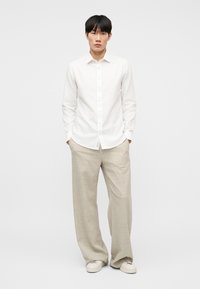 Άνδρας που στέκεται με τα χέρια στις τσέπες, φοράει λευκό πουκάμισο με κουμπιά, μπεζ παντελόνι φαρδύ πόδι και λευκά αθλητικά παπούτσια σε απλό φόντο.